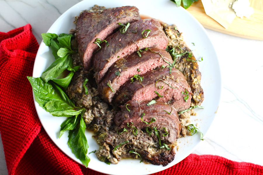 Stuffed Flank Steak cooked and cut on white plate with stuffing oozing out and basil on side. In the stuffing you get mushrooms, onions, and garlic sauteed together. Then they are mixed with fresh parsley and basil, then parmesan, and goat cheese! Roll it all up in Flank Steak and roast for an amazing dinner.