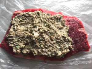 Raw flank steak with Mushroom stuffing spread on top for the Stuffed Flank Steak recipe. it is oozing with flavor! First, you get mushrooms, onions, and garlic sauteed together. Then they are mixed with fresh parsley and basil, then parmesan, and goat cheese! Roll it all up in Flank Steak and roast for an amazing dinner.