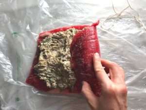 Raw flank steak with Mushroom stuffing spread on top and hand starting to roll the steak from the right side. This Stuffed Flank Steak recipe is oozing with flavor! First, you get mushrooms, onions, and garlic sauteed together. Then they are mixed with fresh parsley and basil, then parmesan, and goat cheese! Roll it all up in Flank Steak and roast for an amazing dinner.