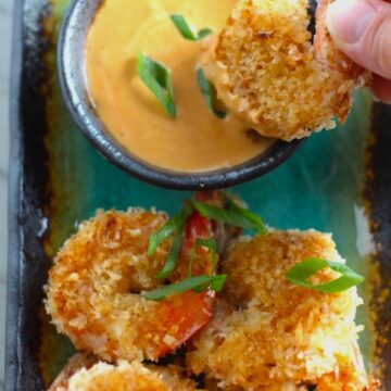 Baked Coconut Shrimp on plate with hand dipping one into Sriracha Sauce. Crunchy and utterly irresistible, this 30-Minute Baked Coconut Shrimp recipe is one that you have got to try!  They are baked, not fried, so they are EASY to cook and HEALTHIER for you too! 