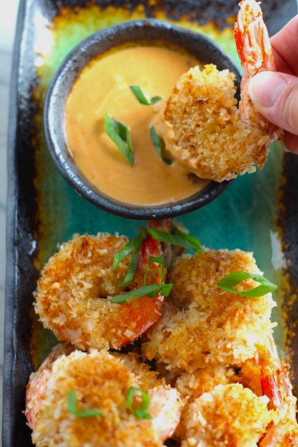 Baked Coconut Shrimp on plate with hand dipping one into Sriracha Sauce. Crunchy and utterly irresistible, this 30-Minute Baked Coconut Shrimp recipe is one that you have got to try! They are baked, not fried, so they are EASY to cook and HEALTHIER for you too!