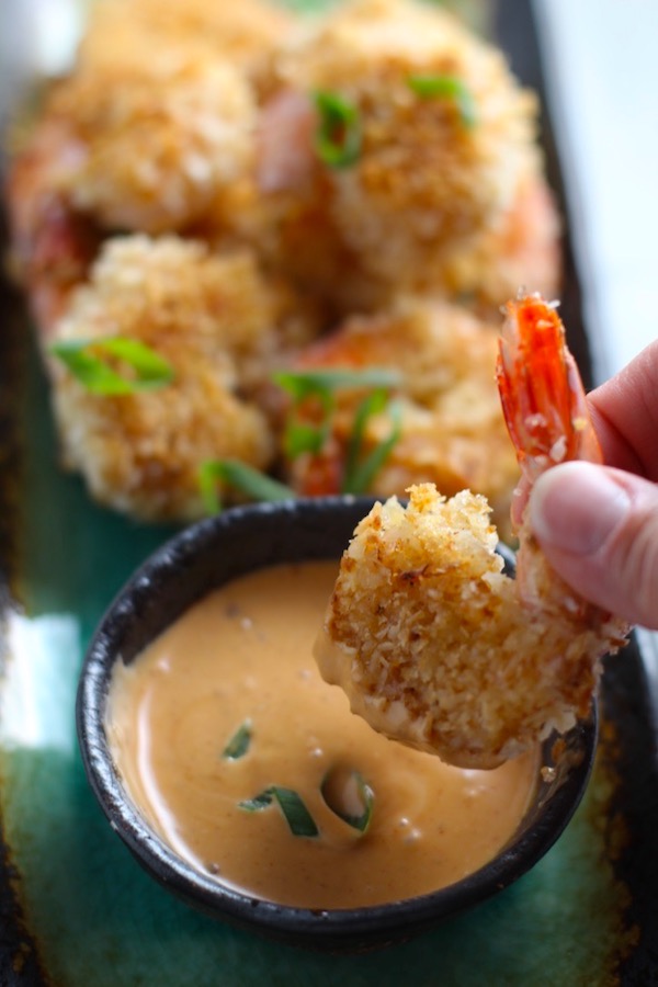 Baked Coconut Shrimp on plate with hand dipping one into Sriracha Sauce. Crunchy and utterly irresistible, this 30-Minute Baked Coconut Shrimp recipe is one that you have got to try! They are baked, not fried, so they are EASY to cook and HEALTHIER for you too!