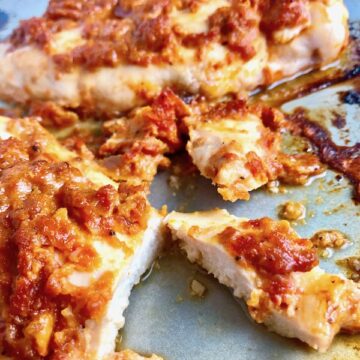 Sun Dried Tomato Chicken cooked on a pan with a piece cut. It's slightly sweet, slightly tart and tangy, and hugely delicious!  When Sun dried tomatoes are rehydrated and blended, they turn into a thick and lucious sauce that coats the chicken keeping it moist and juicy when it simply bakes in the oven.  It's a perfect easy weeknight dinner that you can prepare ahead and cook quickly! #chicken #chickenrecipes #easydinners #dinnerrecipes