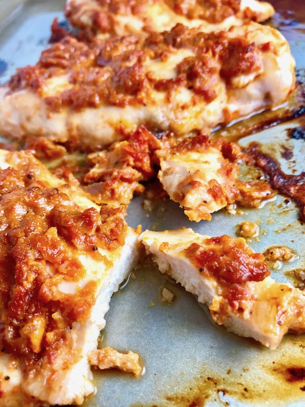 Sun Dried Tomato Chicken cooked on a pan with a piece cut. It's slightly sweet, slightly tart and tangy, and hugely delicious! When Sun dried tomatoes are rehydrated and blended, they turn into a thick and lucious sauce that coats the chicken keeping it moist and juicy when it simply bakes in the oven. It's a perfect easy weeknight dinner that you can prepare ahead and cook quickly! #chicken #chickenrecipes #easydinners #dinnerrecipes