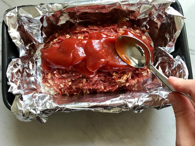 Spoon spreading ketchup on top of raw mexican meatloaf.