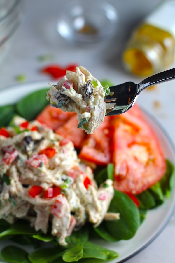 Fork with bite of Curry Chicken Salad on spinach with tomato slices. It has so many fantastic flavors and textures! The chicken breast is simply roasted and mixed with a creamy sweet and savory curry dressing. Red pepper gives you a fresh crunch, scallion gives a savory bite, and raisins give a burst of sweet. #chicken #chickenrecipes #chickensalad #currychicken #mealprep #lunch #healthyrecipes