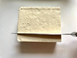 Knife slicing Extra Firm Tofu for Roasted Tofu Croutons. These are seasoned and toasted for the perfect addition to any salad, pasta, rice, or soup! They get a roasted, almost nutty flavor with a salty and smoky seasoned crust with a chewy bite. Super easy to make, low carb and great source of protein! #croutons #salad #tofu #tofurecipes #vegetarian #plantbased #paleo