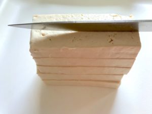 Knife slicing Extra Firm Tofu for Roasted Tofu Croutons. These are seasoned and toasted for the perfect addition to any salad, pasta, rice, or soup! They get a roasted, almost nutty flavor with a salty and smoky seasoned crust with a chewy bite. Super easy to make, low carb and great source of protein! #croutons #salad #tofu #tofurecipes #vegetarian #plantbased #paleo