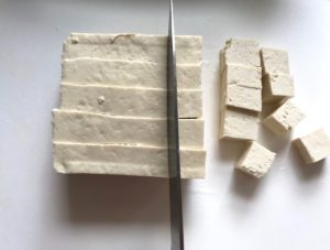 Knife slicing Extra Firm Tofu into pieces for Roasted Tofu Croutons. These are seasoned and toasted for the perfect addition to any salad, pasta, rice, or soup! They get a roasted, almost nutty flavor with a salty and smoky seasoned crust with a chewy bite. Super easy to make, low carb and great source of protein! #croutons #salad #tofu #tofurecipes #vegetarian #plantbased #paleo