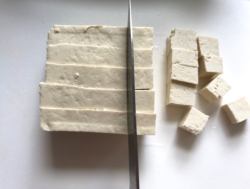 Knife slicing Extra Firm Tofu into pieces for Roasted Tofu Croutons. These are seasoned and toasted for the perfect addition to any salad, pasta, rice, or soup! They get a roasted, almost nutty flavor with a salty and smoky seasoned crust with a chewy bite. Super easy to make, low carb and great source of protein! #croutons #salad #tofu #tofurecipes #vegetarian #plantbased #paleo