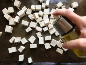 Hand spraying tofu pieces with olive oil for Roasted Tofu Croutons. These are seasoned and toasted for the perfect addition to any salad, pasta, rice, or soup! They get a roasted, almost nutty flavor with a salty and smoky seasoned crust with a chewy bite. Super easy to make, low carb and great source of protein! #croutons #salad #tofu #tofurecipes #vegetarian #plantbased #paleo