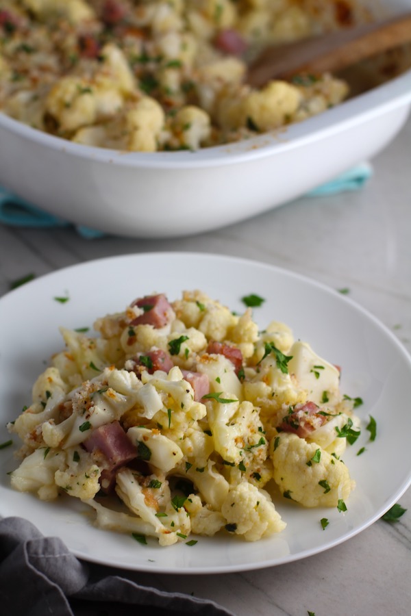 Gluten Free Cauliflower Cheese and Ham Bake on plate with casserole dish in background. It layers in nutty and salty Gruyere Cheese, with a creamy Vegetable broth based sauce with garlic and parsley. Then the ham and cauliflower are folded into all of this goodness and it's baked in the oven until it all becomes one knock-your-socks-off dish! #cauliflower #lowcarb #glutenfree #casserole #onepot #familydinner #mealprep #easydinner #dinner #healthydinner #healthyfood #healthyrecipes