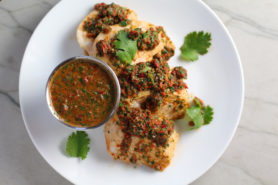 Chicken Chimichurri Rojo on a plate with cup of chimichurri. It is broiled or grilled Chicken topped with a cool, fresh, tangy, and incredibly full-flavored! The Rojo comes from the addition of red pepper. All of the ingredients simply get blended in a food processor so it could not be easier! #chicken #chickenrecipes #chimichurri #easychicken #easydinners #dinner