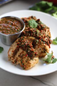 Chicken Chimichurri Rojo on a plate with cup of chimichurri. It is broiled or grilled Chicken topped with a cool, fresh, tangy, and incredibly full-flavored! The Rojo comes from the addition of red pepper. All of the ingredients simply get blended in a food processor so it could not be easier! #chicken #chickenrecipes #chimichurri #easychicken #easydinners #dinner