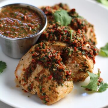 Chicken Chimichurri Rojo on a plate with cup of chimichurri. It is broiled or grilled Chicken topped with a cool, fresh, tangy, and incredibly full-flavored!  The Rojo comes from the addition of red pepper.  All of the ingredients simply get blended in a food processor so it could not be easier!  #chicken #chickenrecipes #chimichurri #easychicken #easydinners #dinner
