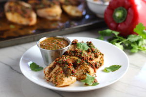 Chicken Chimichurri Rojo on a plate with cup of chimichurri. It is broiled or grilled Chicken topped with a cool, fresh, tangy, and incredibly full-flavored! The Rojo comes from the addition of red pepper. All of the ingredients simply get blended in a food processor so it could not be easier! #chicken #chickenrecipes #chimichurri #easychicken #easydinners #dinner