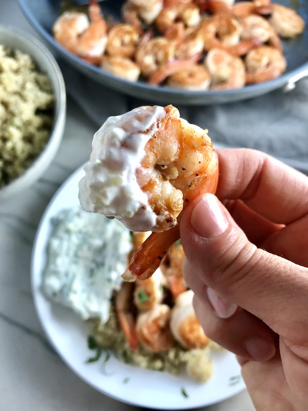 Hand holding Garlic and Lime Shrimp dipped in Tzatziki Sauce with plates in background. The Shrimp is simply sauteed with minced garlic, olive oil, and lime for big bold flavors. The Tzatziki Sauce is bright and cool with creamy yogurt with fresh cucumber, lime, garlic, and scallion. #shrimp #easydinner #dinner #seafoodrecipes #shrimprecipes