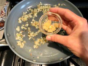 Adding garlic to onions cooking in pan for Artichoke Spinach Stuffed Zucchini Recipe. Each fantastic bite gives you creamy artichoke, nutty cheesy Parmesan, spinach, and zucchini. Prepare entirely ahead, then bake 20 minutes and enjoy! #vegetarian #zucchini #stuffedzuchini #spinach #artichoke #springrecipes #healthyfood #healthydinner #healthyrecipes #glutenfree