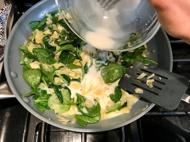 Adding cornstarch slurry to spinach and artichokes in pan for Artichoke Spinach Stuffed Zucchini Recipe. Each fantastic bite gives you creamy artichoke, nutty cheesy Parmesan, spinach, and zucchini. Prepare entirely ahead, then bake 20 minutes and enjoy! #vegetarian #zucchini #stuffedzuchini #spinach #artichoke #springrecipes #healthyfood #healthydinner #healthyrecipes #glutenfree