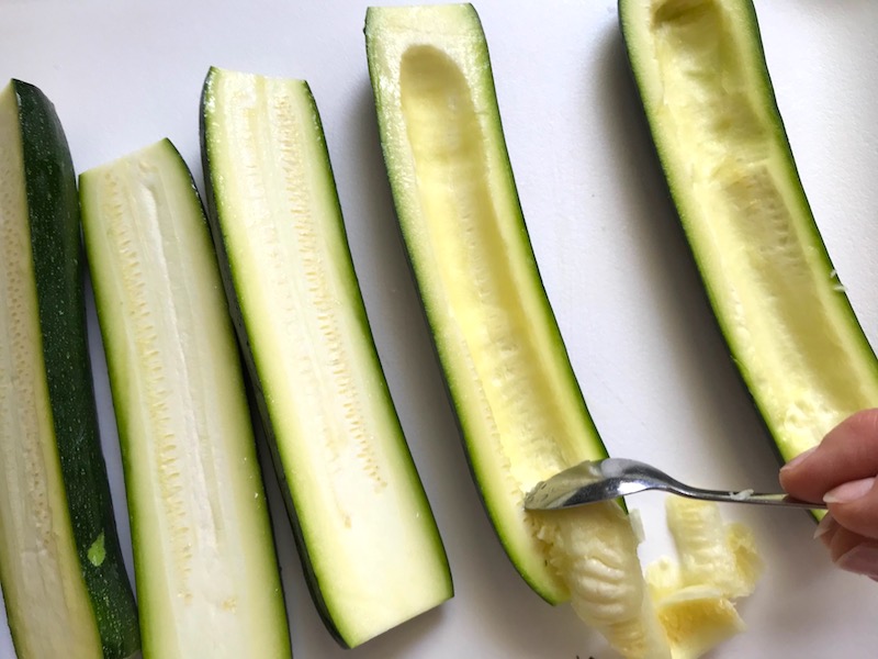 Spoon scooping seeds of zucchini half for Artichoke Spinach Stuffed Zucchini Recipe. Each fantastic bite gives you creamy artichoke, nutty cheesy Parmesan, spinach, and zucchini. Prepare entirely ahead, then bake 20 minutes and enjoy! #vegetarian #zucchini #stuffedzuchini #spinach #artichoke #springrecipes #healthyfood #healthydinner #healthyrecipes #glutenfree