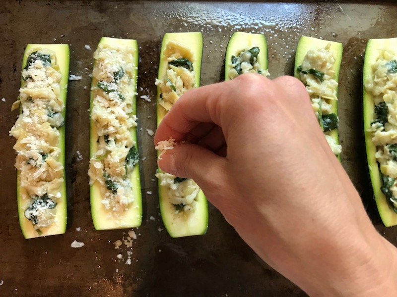 Sprinkling parmesan on stuffed zucchini halves for Artichoke Spinach Stuffed Zucchini Recipe. Each fantastic bite gives you creamy artichoke, nutty cheesy Parmesan, spinach, and zucchini. Prepare entirely ahead, then bake 20 minutes and enjoy! #vegetarian #zucchini #stuffedzuchini #spinach #artichoke #springrecipes #healthyfood #healthydinner #healthyrecipes #glutenfree