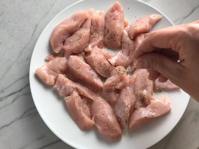 Hand sprinkling seasoning on raw chicken pieces for Chicken and Cherry Tomato Pasta with basil and parmesan. It's easy and so delicious! #pasta #tomatoes #easydinner #dinner #easyrecipes #healthydinner #chicken