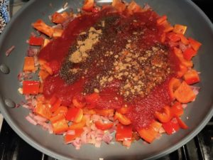 Spices, tomato sauce, diced onion, red pepper, and garlic in skillet cooking for Skillet Eggs in Spicy Tomato Pepper Sauce with Manchego Cheese. With a smoky, spicy, thick, and luscious red pepper tomato sauce. #eggs #huevosrancheros #tomatosauce #easydinner #brunch #shakshuka