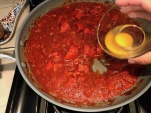 Hand pouring raw egg into hole in tomato sauce in pan for Skillet Eggs in Spicy Tomato Pepper Sauce with Manchego Cheese. With a smoky, spicy, thick, and luscious red pepper tomato sauce. #eggs #huevosrancheros #tomatosauce #easydinner #brunch #shakshuka