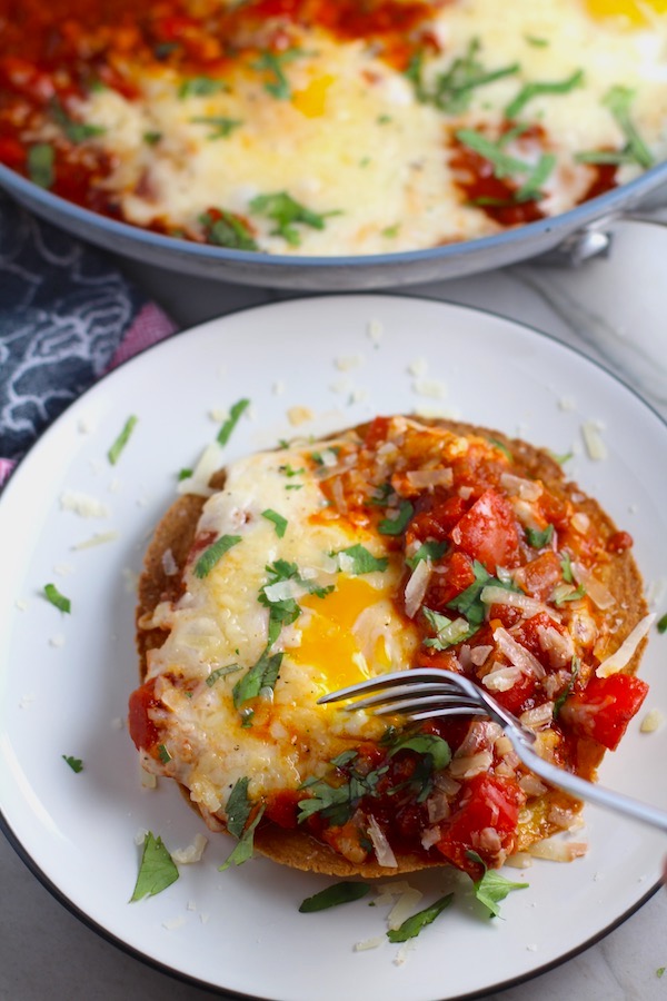 Skillet Eggs in Spicy Tomato Pepper Sauce with Manchego Cheese served over a crispy corn tortilla with skillet in back. With a smoky, spicy, thick, and luscious red pepper tomato sauce. #eggs #huevosrancheros #tomatosauce #easydinner #brunch #shakshuka