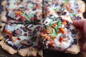 Hand Holding piece of Flatbread with crispy gluten free crust, salty and briny Olive Tapenade, creamy mozzarella, earthy and meaty mushrooms, peppery pancetta, and fresh basil. #flatbread #pizza #olives #tapenade #mediterranean #easydinner #dinner