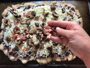 Hand putting pancetta and mushrooms on cheese on flatbread for Olive & Pancetta Flatbread with mozzarella, mushrooms, and fresh basil. #flatbread #pizza #olives #tapenade #mediterranean #easydinner #dinner