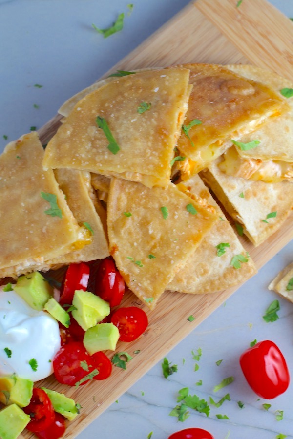 Chicken Corn Tortilla Quesadilla triangles on platter with avocado, tomato, & sour cream. They have crispy edges, a soft gooey center, and are mouthwatering flavor. They have hearty shredded chicken flavored with warm and smoky Mexican spices. Then shredded Pepper Jack cheese and Cheddar is layered to get melty and oozy and delicious. It's an irresistible new easy dinner or appetizer idea. #quesadillas #mexicanfood #enchiladas #familydinner #chicken