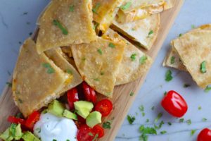 Chicken Corn Tortilla Quesadilla triangles on platter with avocado, tomato, & sour cream. They have crispy edges, a soft gooey center, and are mouthwatering flavor. They have hearty shredded chicken flavored with warm and smoky Mexican spices. Then shredded Pepper Jack cheese and Cheddar is layered to get melty and oozy and delicious. It's an irresistible new easy dinner or appetizer idea. #quesadillas #mexicanfood #enchiladas #familydinner #chicken