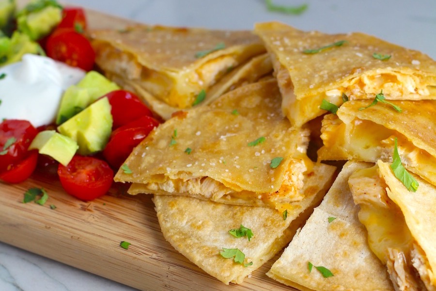 Chicken Corn Tortilla Quesadilla triangles on platter with avocado, tomato, & sour cream. They have crispy edges, a soft gooey center, and are mouthwatering flavor. They have hearty shredded chicken flavored with warm and smoky Mexican spices. Then shredded Pepper Jack cheese and Cheddar is layered to get melty and oozy and delicious. It's an irresistible new easy dinner or appetizer idea. #quesadillas #mexicanfood #enchiladas #familydinner #chicken