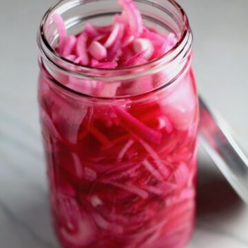 Perfectly Pink Pickled Onions in a mason jar with lid off. They are tart, tangy, sweet, and savory.  They go on just about anything and are so unbelievably delicious, they can make a shoe taste good!  Best part, you can make a ton of them ahead!