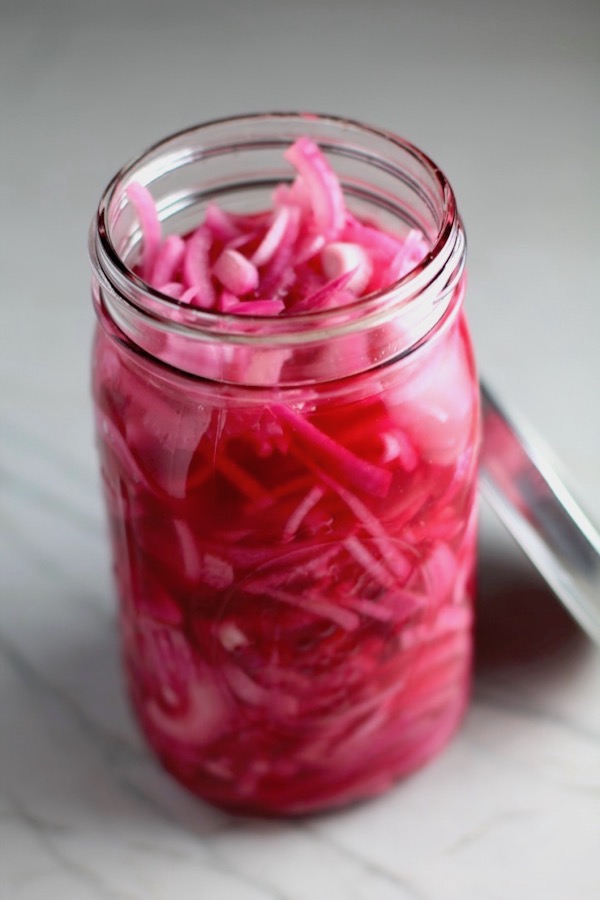 Perfectly Pink Pickled Onions in a mason jar with lid off. They are tart, tangy, sweet, and savory. They go on just about anything and are so unbelievably delicious, they can make a shoe taste good! Best part, you can make a ton of them ahead!
