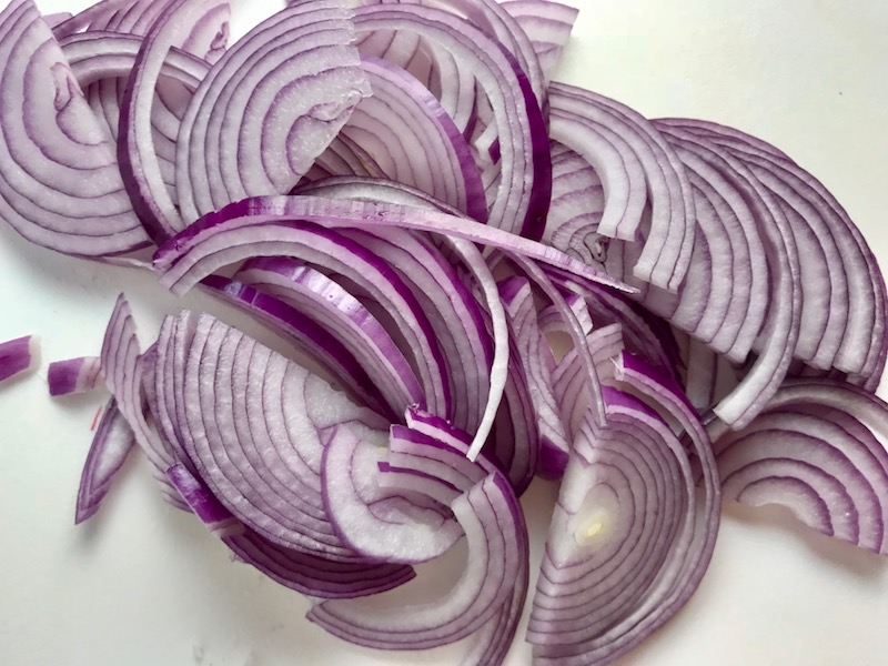Slices of red onion for Pickled Onions in a bowl soaking. They are tart, tangy, sweet, and savory. They go on just about anything and are so unbelievably delicious, they can make a shoe taste good! Best part, you can make a ton of them ahead!
