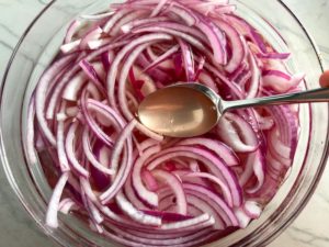 Slices of red onion with pickling liquid in bowl for Pickled Onions in a bowl soaking. They are tart, tangy, sweet, and savory. They go on just about anything and are so unbelievably delicious, they can make a shoe taste good! Best part, you can make a ton of them ahead!