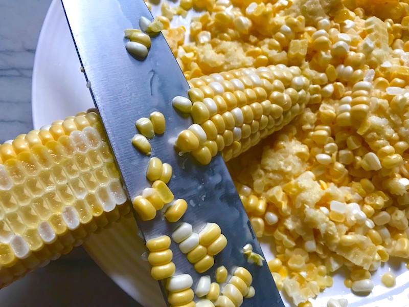 Knife cutting corn kernels off cob for Chilled Golden Corn Soup with Turmeric. Scallions on top for garnish. It's thick, creamy, silky and delicious. The entire family will love this easy stovetop corn soup! #summerfood #cornsoup #corn #vegetarian #vegetablerecipes #sides