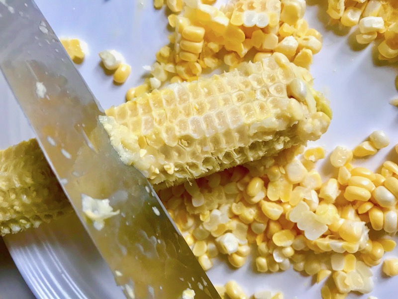 Back of Knife scraping the 'cream' from a cob of corn for Gluten Free Corn Chowder with Turmeric. Scallions on top for garnish.