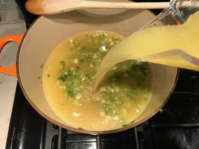 Pouring broth over sliced scallions in a soup pot for Chilled Golden Corn Soup with Turmeric. Scallions on top for garnish. It's thick, creamy, silky and delicious. The entire family will love this easy stovetop corn soup! #summerfood #cornsoup #corn #vegetarian #vegetablerecipes #sides
