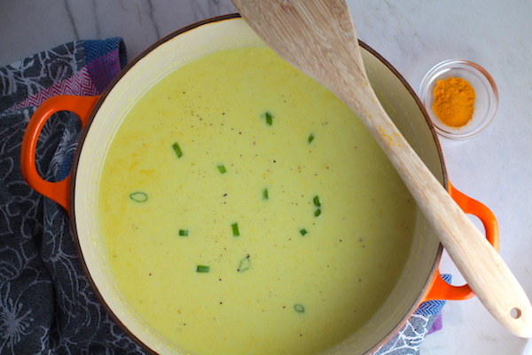 Chilled Golden Corn Soup in a pot with a wooden spoon on side and turmeric on counter. Scallions on top for garnish. It's thick, creamy, silky and delicious. The entire family will love this easy stovetop corn soup! #summerfood #cornsoup #corn #vegetarian #vegetablerecipes #sides