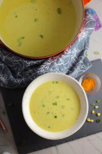 Chilled Golden Corn Soup in a bowl with pot of soup behind and turmeric spice in a bowl on the side. Scallions on top for garnish. It's thick, creamy, silky and delicious. The entire family will love this easy stovetop corn soup! #summerfood #cornsoup #corn #vegetarian #vegetablerecipes #sides