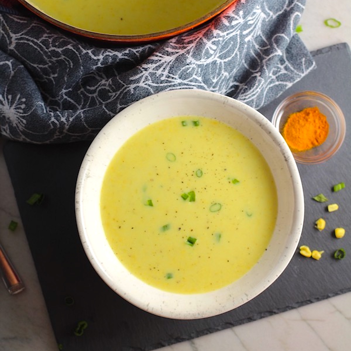 Gluten free Corn Chowder in a bowl scallions on top as garnish, a small bowl of turmeric next to it and a pot of the chowder in the background.
