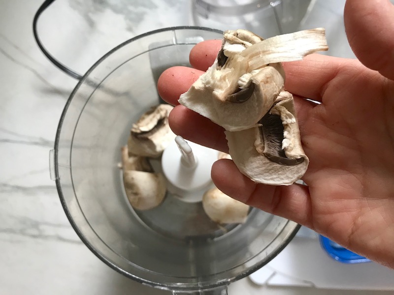Hand dropping mushrooms into processor for Thick & Creamy Healthy Mushroom Soup. The soup has earthy mushrooms, aeromatic onions, flavorful oregano, and bright chives.