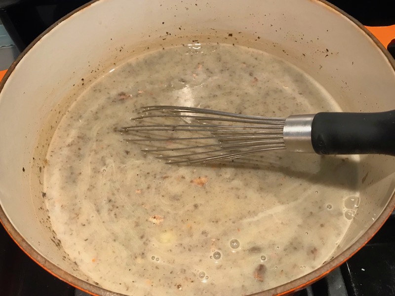 Whisk stirring Creamy Healthy Mushroom Soup in a pot filled with earthy mushrooms, aeromatic onions, flavorful oregano, and bright chives.