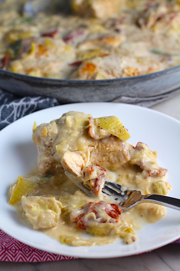 Close up of Artichoke Chicken with sun dried tomatoes on plate with fork holding a bite and pan in background. #skilletchicken #skilletdinner #chicken #easydinner #familydinner #italian #tuscanchicken