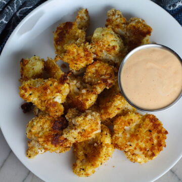 Baked Coconut Crispy Cauliflower Bites on plate with Sriracha Mayo dipping sauce. They have a slightly sweet and salty crunch outside from the shredded coconut and panko mixture. The inside is soft and creamy. Dip in the Sriracha Mayo.