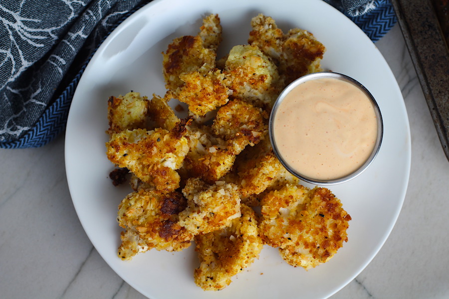 Baked Coconut Cauliflower Bites on plate with Sriracha Mayo dipping sauce. They have a slightly sweet and salty crunch outside from the shredded coconut and panko mixture. The inside is soft and creamy. Dip in the Sriracha Mayo.
