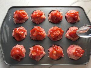 Spoon spreading ketchuo on Raw Mini Meatloaf Muffins in muffin tin. They have only 5 simple ingredients, cook in 30 minutes, and are gluten free.